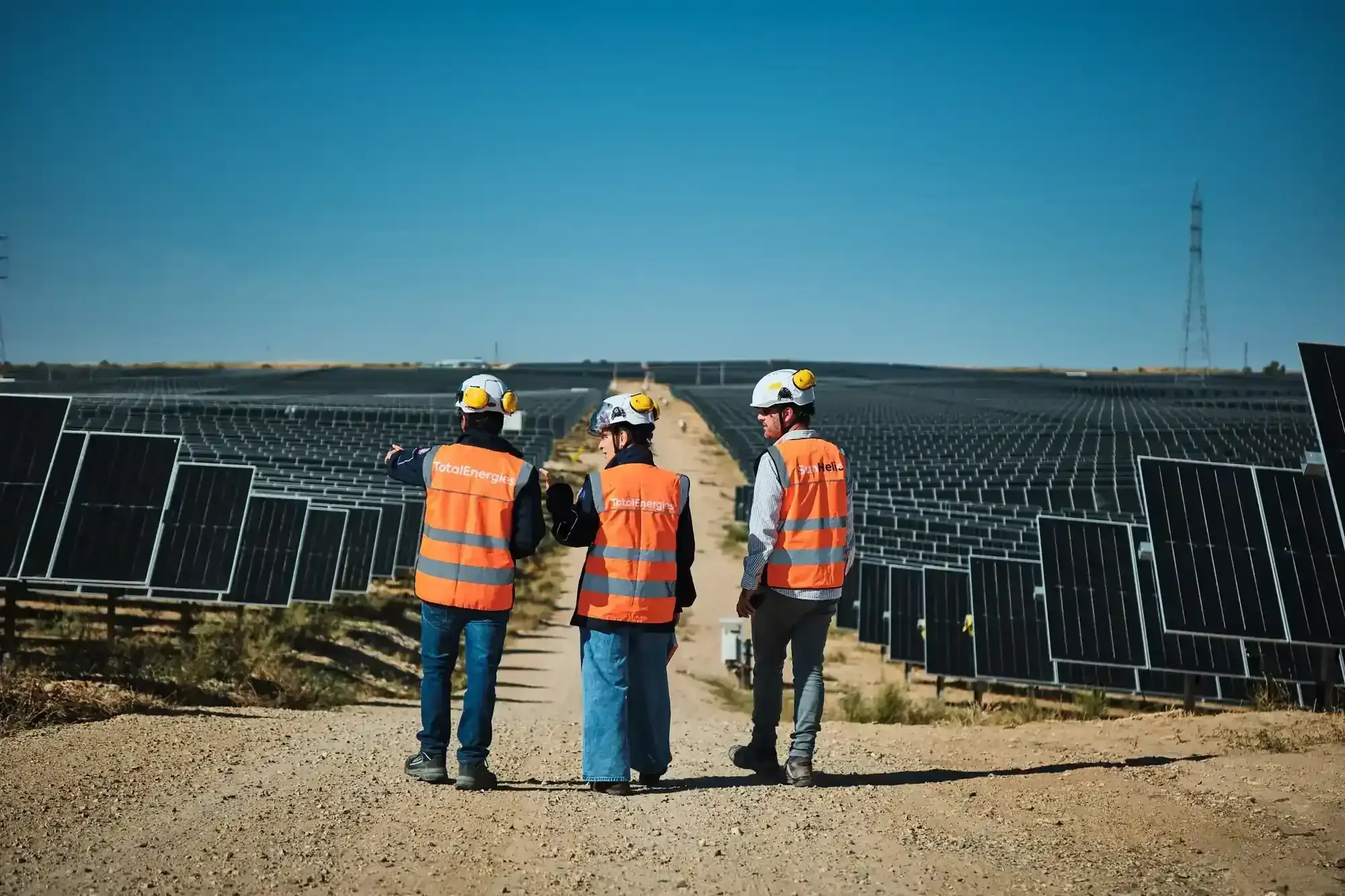 Tres trabajadores vestidos con chaleco naranja de espaldas ante un parque fotovoltaico