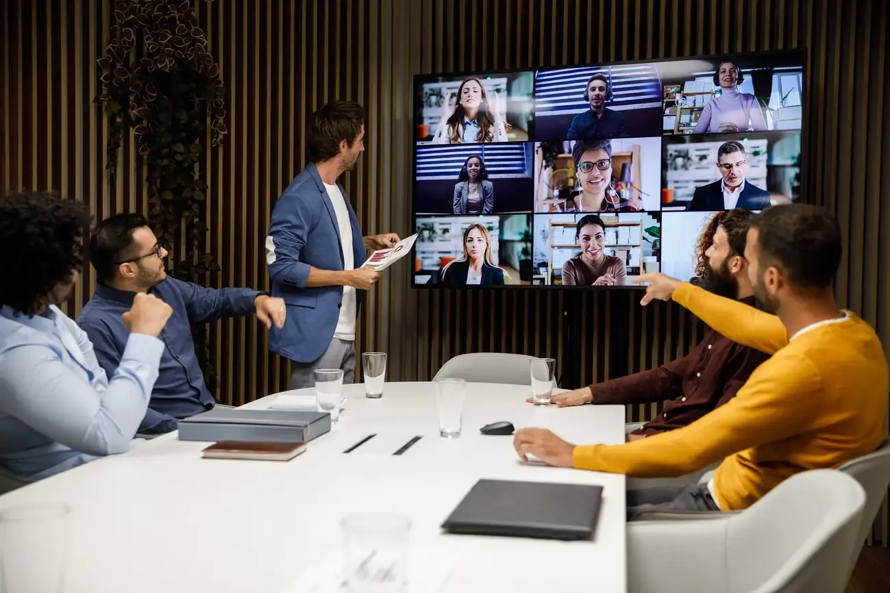 An image of several people in a meeting room having a webianr with others via a large television screen.