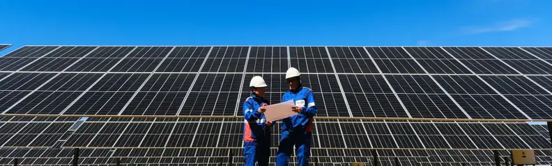 Dos operarios con uniforme de trabajo consultando unos documentos frente a un parque fotovoltaico