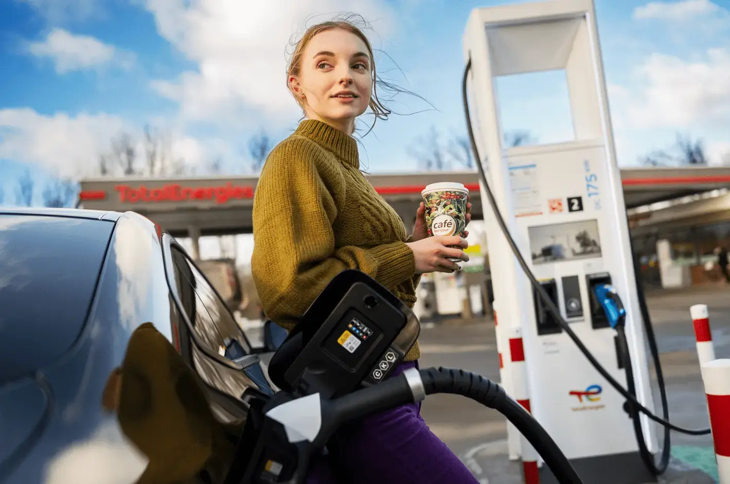 Fahrerin lädt ihr Auto und trinkt Kaffee an TotalEnergies Tankstelle