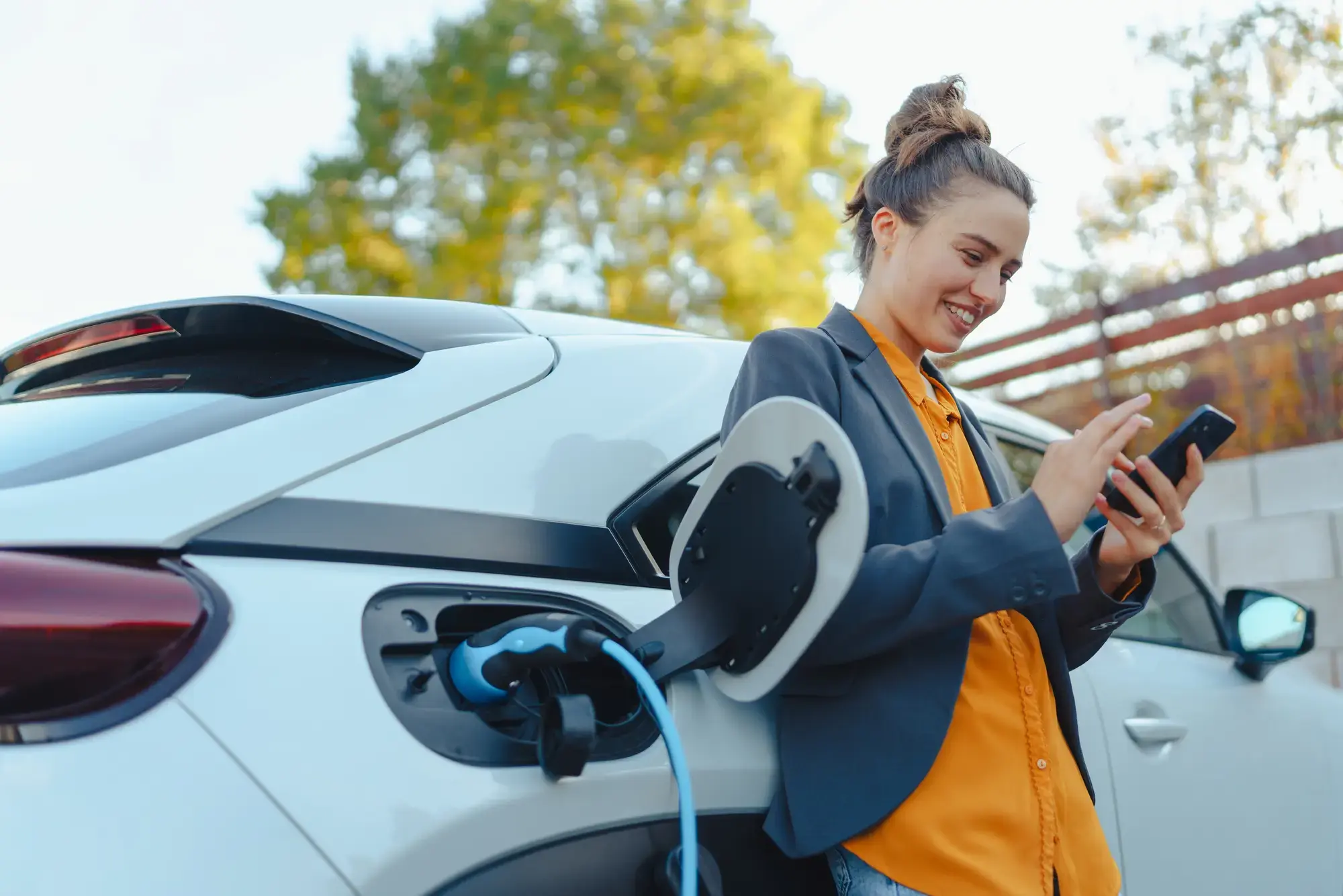 Young woman with smartphone waiting while her electric car charging in home charging station, sustainable and economic transportation concept